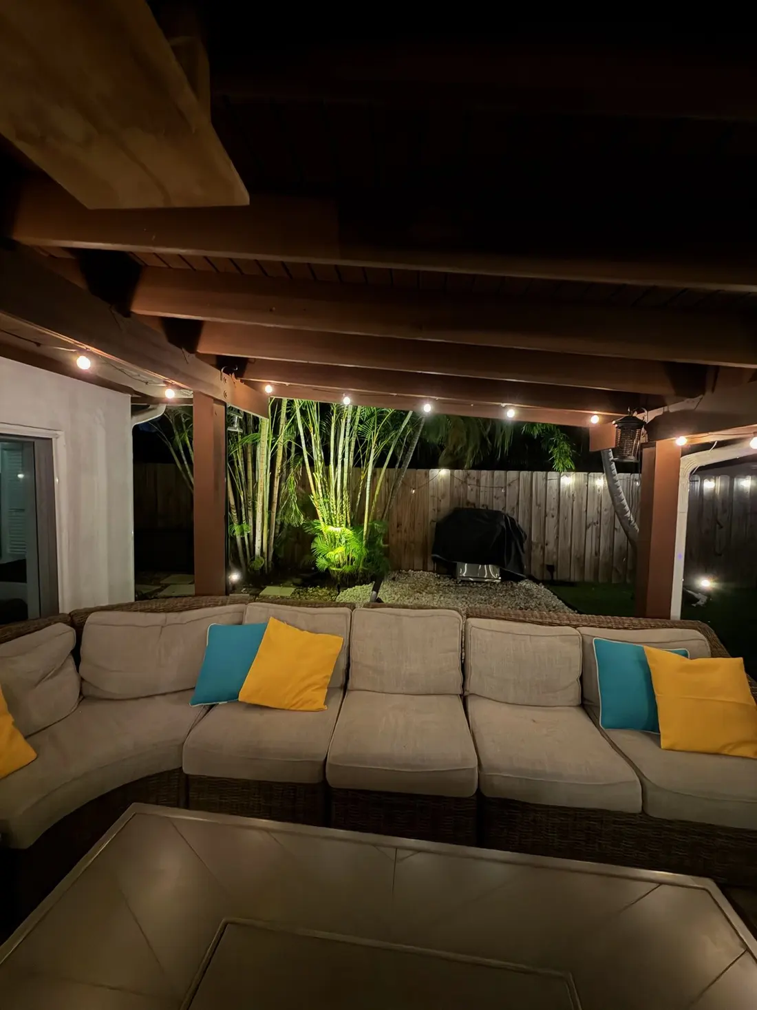 The covered patio sectional with teal and yellow pillows at night, bamboo uplit green behind it
