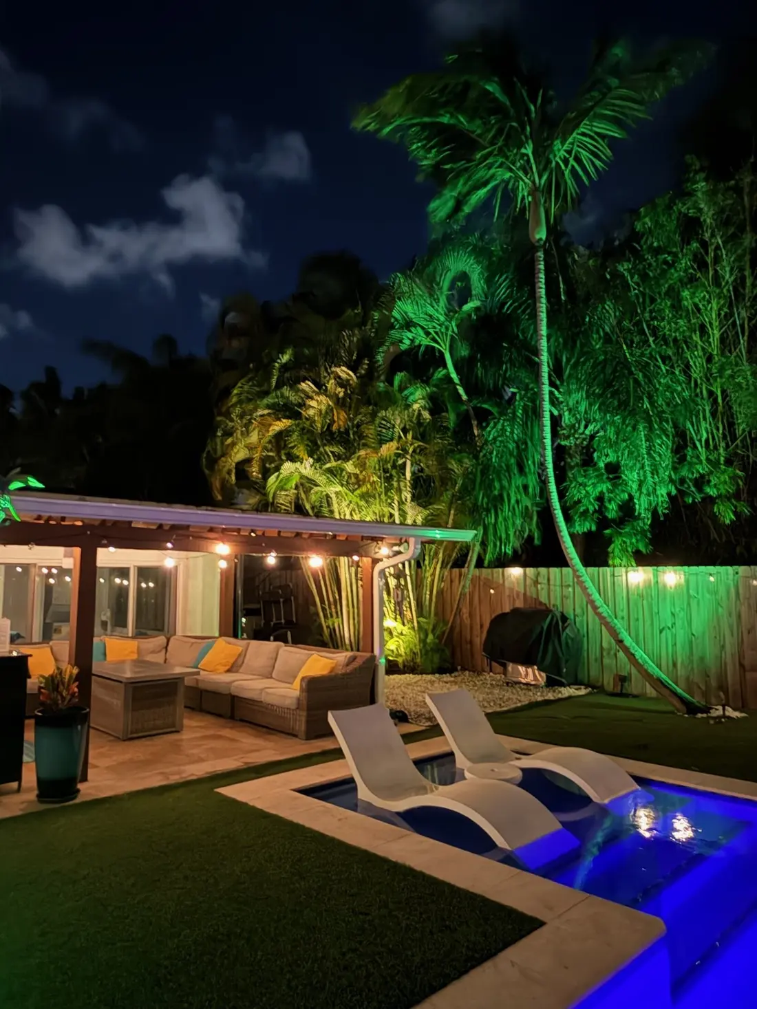 Loungers on the pool's sun shelf at night, the patio lounge and palms uplit green behind