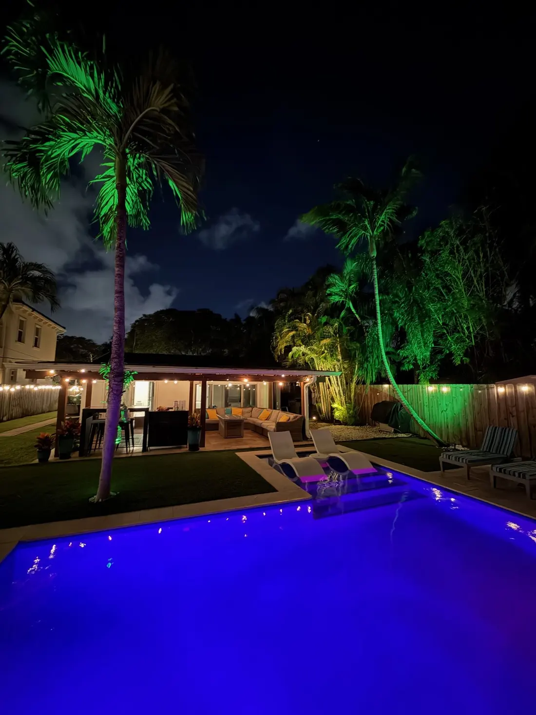 The lit pool and sun-shelf loungers at night, with green-lit palms and the covered patio across the lawn