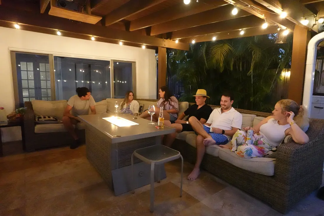 Friends and family talking on the covered patio sectional around the lit fire table, string lights overhead