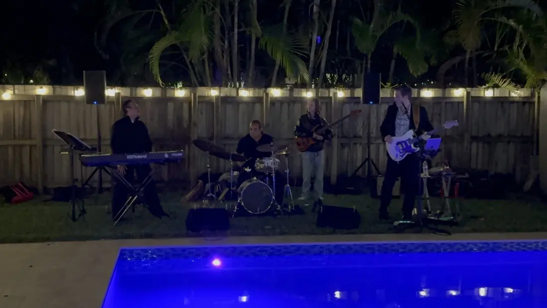 A four-piece band playing beside the lit pool at night, in front of the string-lit backyard fence