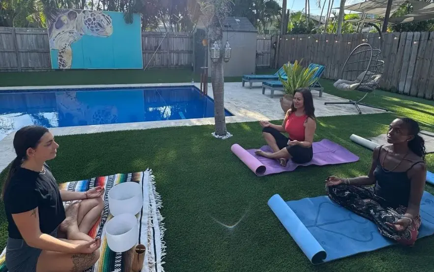 Three women meditating on yoga mats on the lawn beside the pool, with singing bowls, the turtle mural behind them