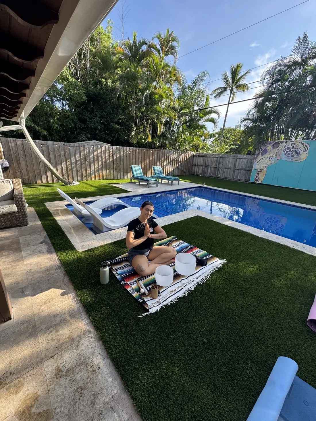 A woman on a striped blanket with singing bowls, doing yoga on the lawn beside the pool