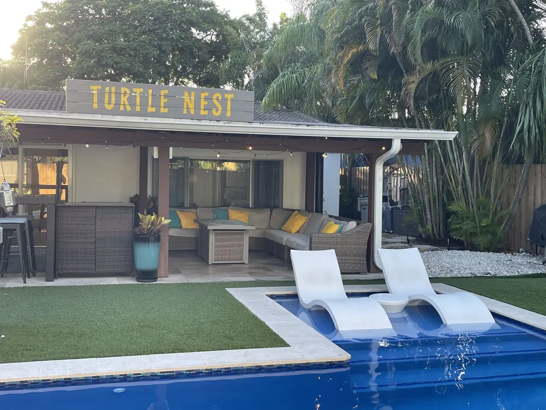 The covered patio under the yellow TURTLE NEST sign: a wicker sectional with yellow and teal pillows, a fire table, a potted croton, and two white loungers on the pool's sun shelf