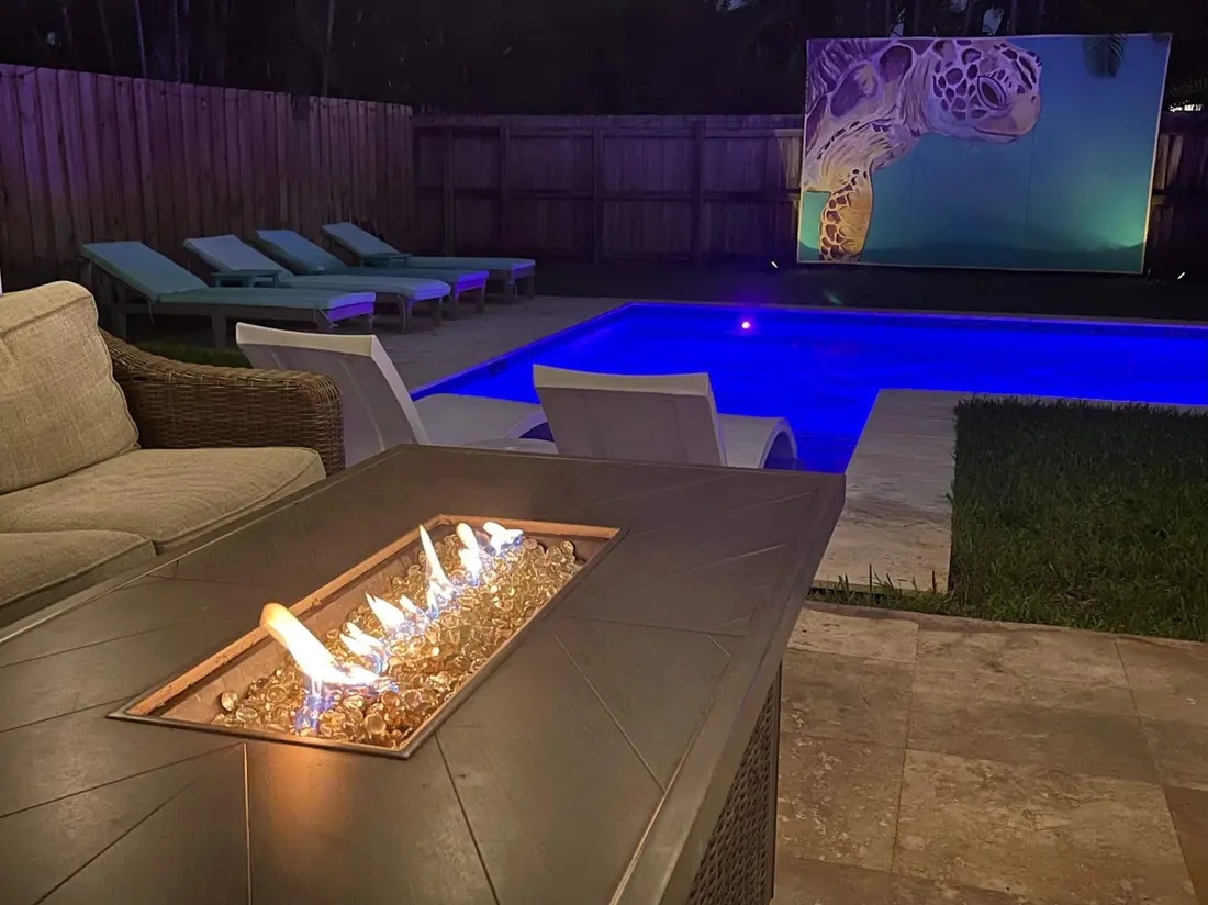 The backyard at night with the fire pit, glowing pool and a large screen