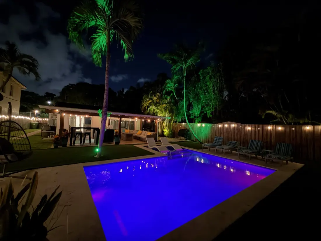 The backyard at night: the pool lit blue, palms uplit green and string lights along the fence