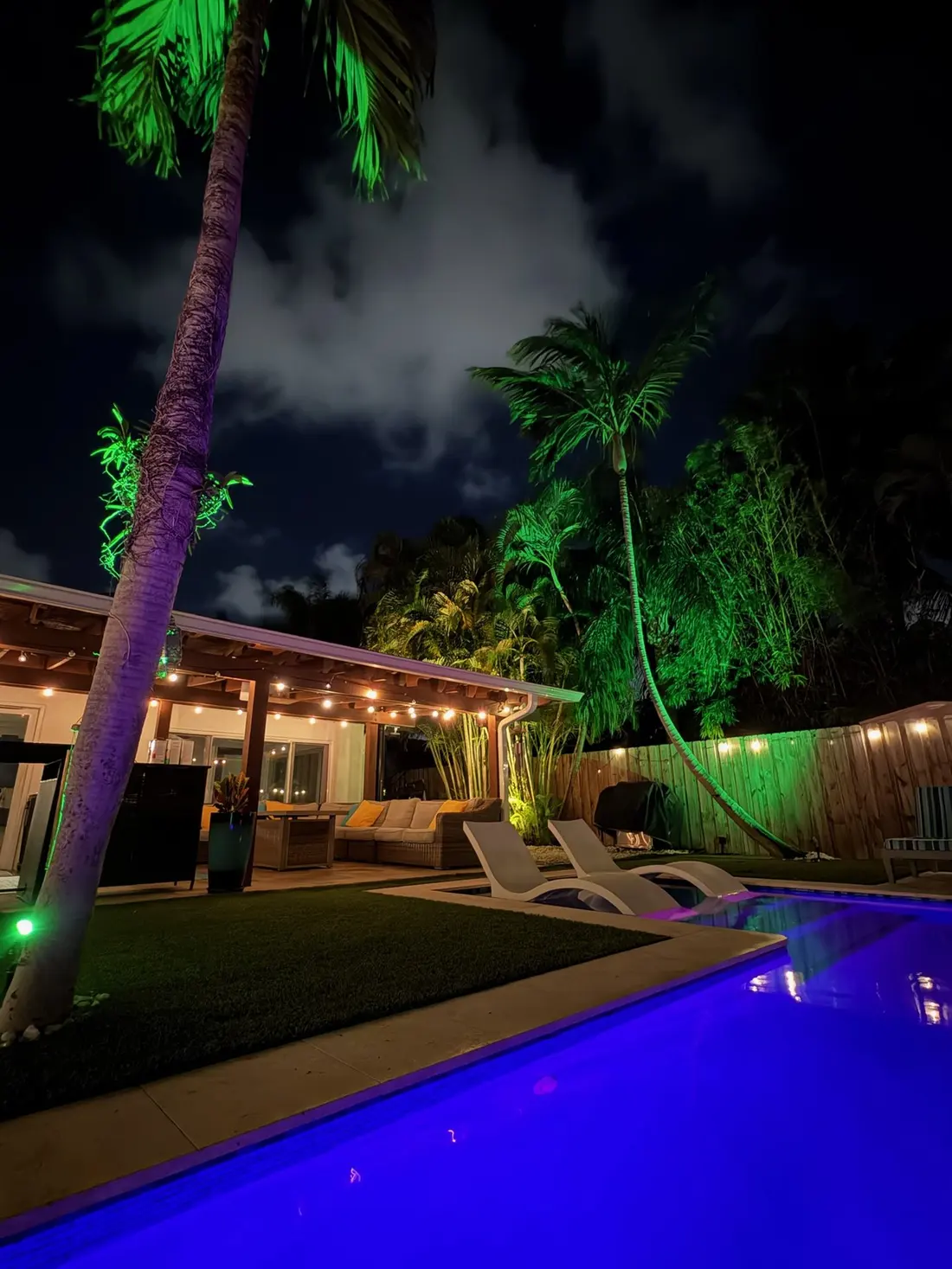 The pool glowing blue at night beside green-lit palms and the string-lit covered patio