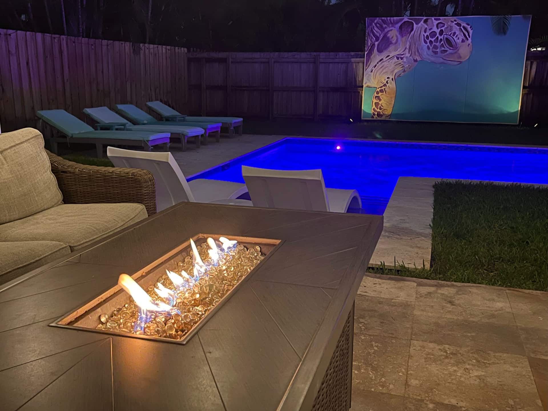 The backyard at night with the fire pit, glowing pool and a large screen