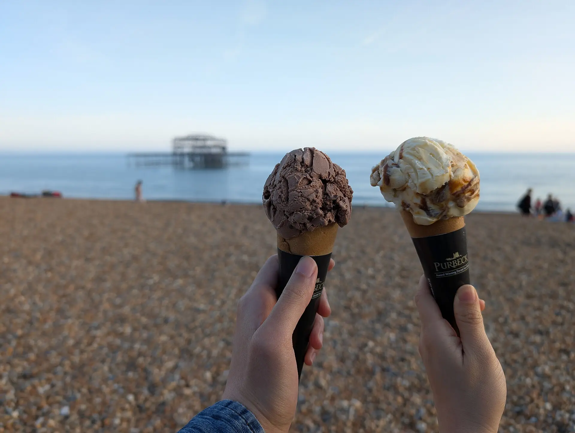 Two ice cream cones held up against a beach and the sea