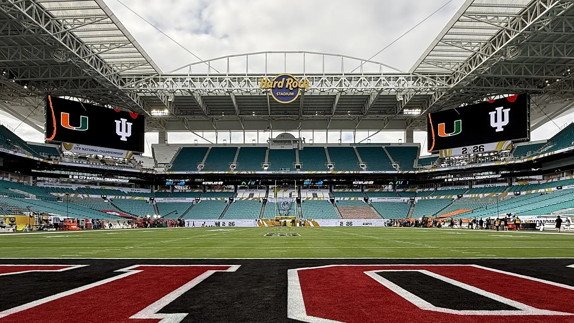 Interior field-level view of Hard Rock Stadium looking across the football field toward empty teal seats, video boards, and the canopy roof