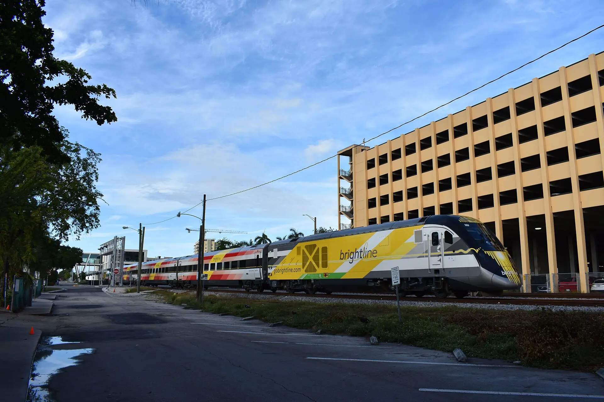A Brightline passenger train travelling along tracks next to a multi-story parking garage and an empty parking lot