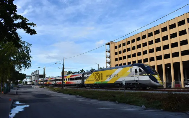 A Brightline passenger train travelling along tracks next to a multi-story parking garage and an empty parking lot
