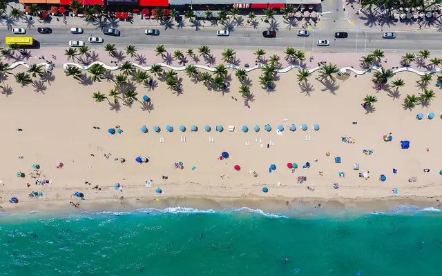Las Olas Oceanside Park: a top-down aerial photograph of Fort Lauderdale Beach showing State Road A1A, the iconic undulating white wave wall, palm trees, sandy beach with umbrellas