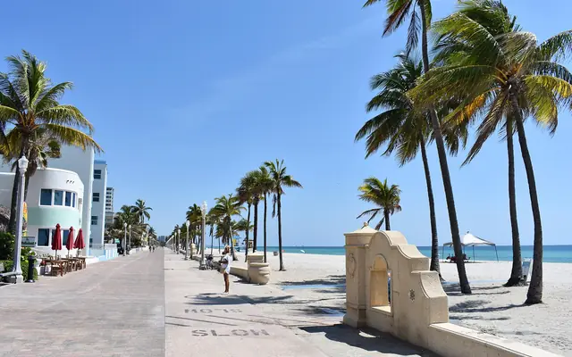 A sunny day view along the paved Hollywood Beach Broadwalk lined with palm trees, seaside buildings, sandy beach, and the ocean