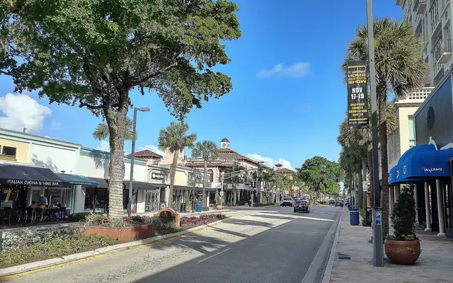 A sunlit street view of Las Olas Boulevard lined with palms, retail storefronts, and restaurant patios