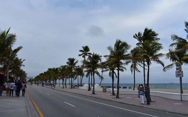 A street-level view looking down State Road A1A along Fort Lauderdale Beach, featuring palm trees, the white wave wall, and pedestrians under an overcast sky