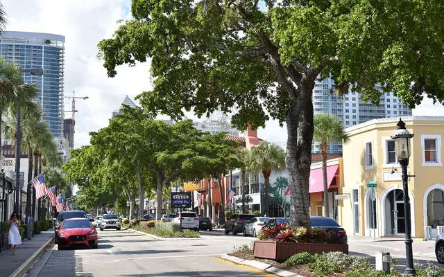 A daytime street view down tree-lined Las Olas Boulevard in Fort Lauderdale, showing shops, cars, and downtown towers in the distance