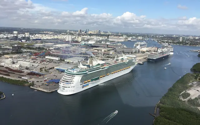 An aerial view of large cruise ships docked and maneuvering through Port Everglades in Fort Lauderdale, with the port facilities and distant skyline visible