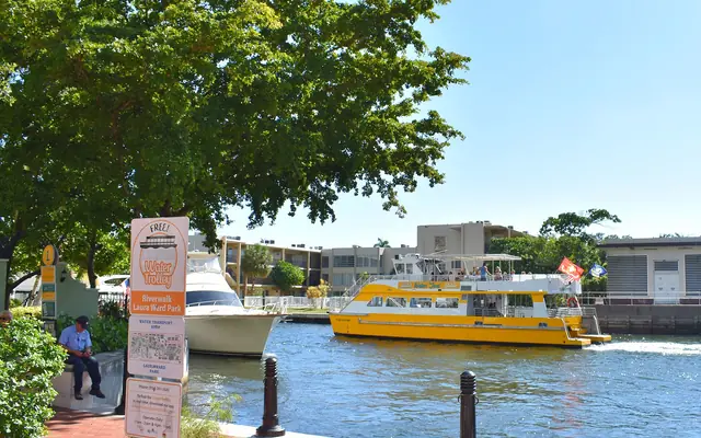 A yellow Fort Lauderdale Water Taxi boat navigates a canal seen from the Riverwalk at Laura Ward Park