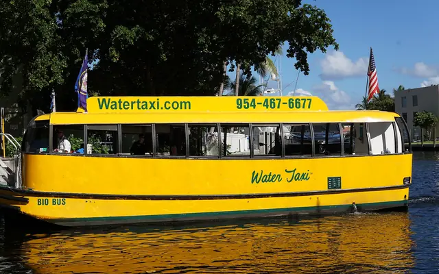 A yellow Fort Lauderdale Water Taxi boat cruising along a waterway with lush green trees and an American flag in the background