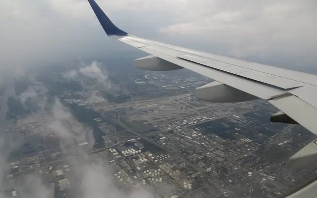 An aerial view through an airplane window showing the aircraft wing above Fort Lauderdale-Hollywood International Airport and Port Everglades under cloudy skies