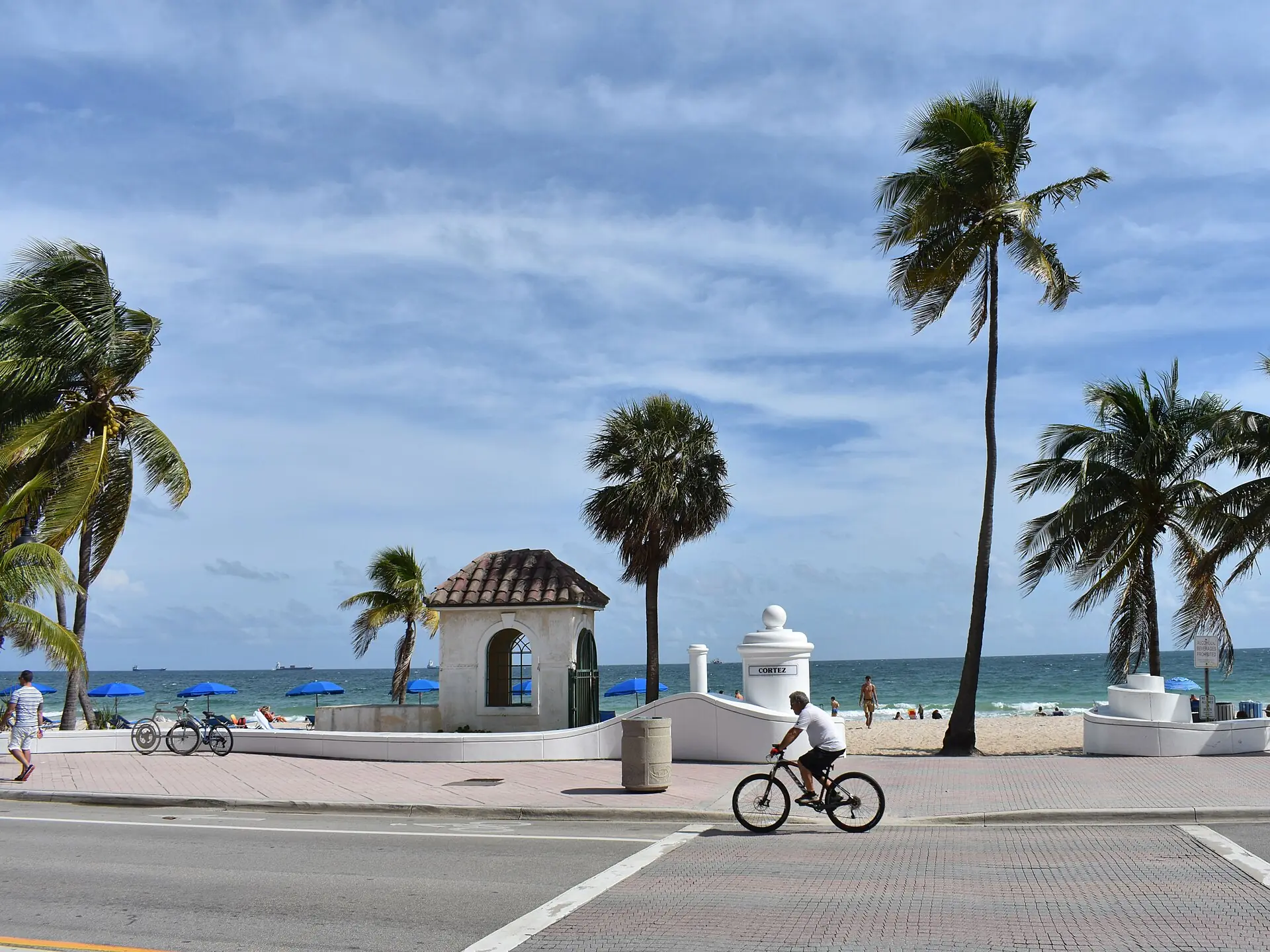 Fort Lauderdale Beach: street-level view across State Road A1A toward the white wave wall, palm trees, and beach at Cortez Street in Fort Lauderdale