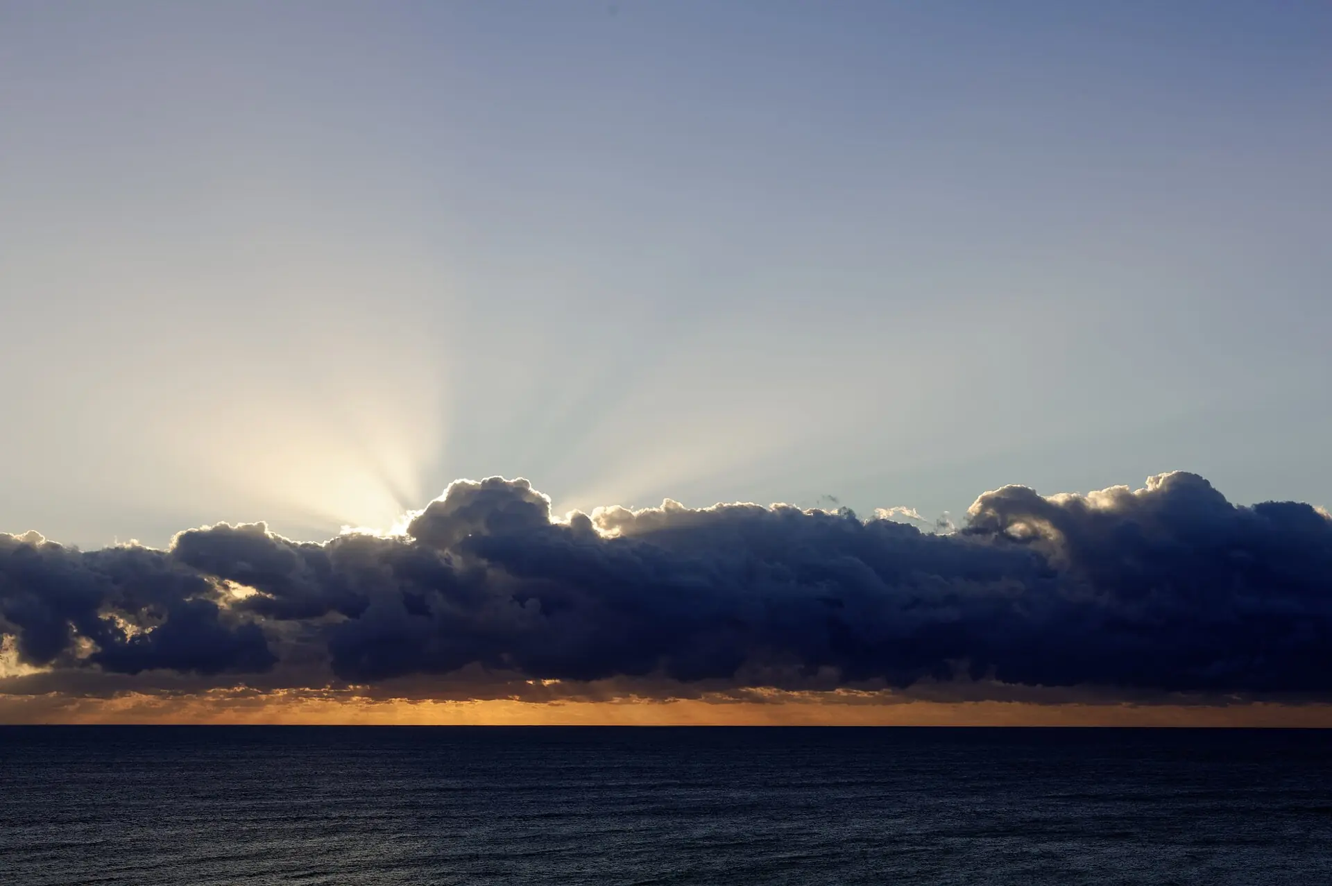 A line of dark clouds over an open ocean with crepuscular sunbeams radiating upward into the sky
