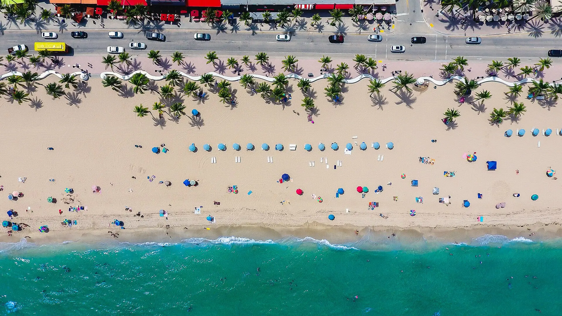 Las Olas Oceanside Park: a top-down aerial photograph of Fort Lauderdale Beach showing State Road A1A, the iconic undulating white wave wall, palm trees, sandy beach with umbrellas