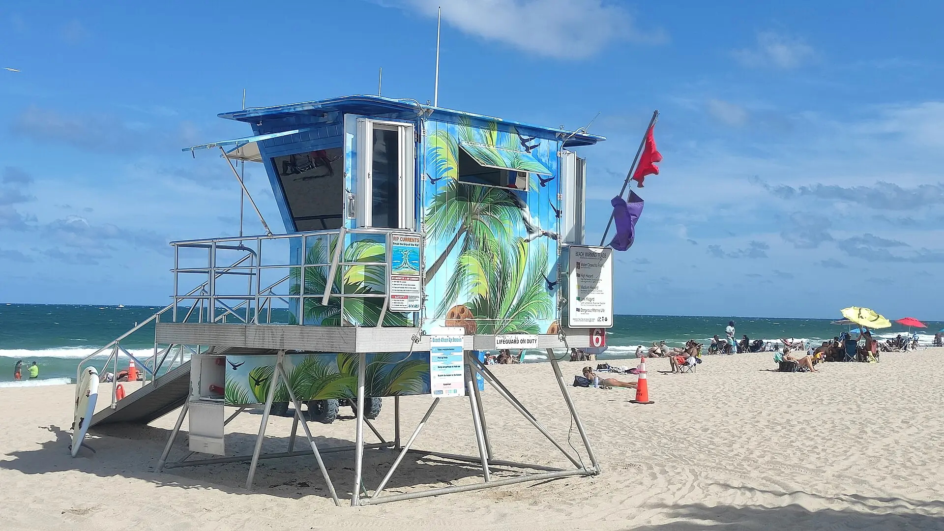 A colorful painted lifeguard tower stands on a sandy beach under a clear blue sky with ocean and beachgoers in the background
