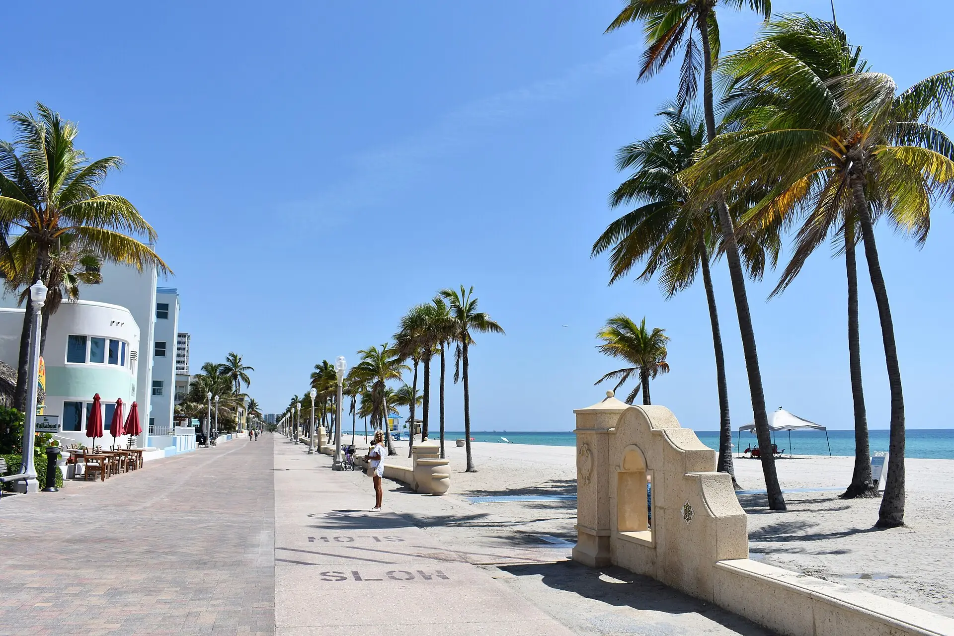 A sunny day view along the paved Hollywood Beach Broadwalk lined with palm trees, seaside buildings, sandy beach, and the ocean