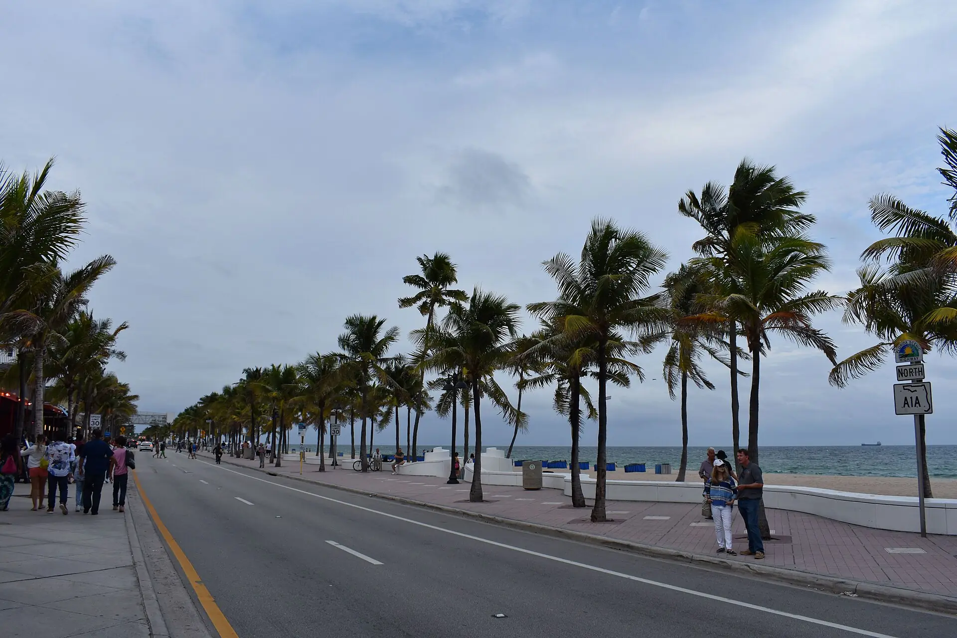 A street-level view looking down State Road A1A along Fort Lauderdale Beach, featuring palm trees, the white wave wall, and pedestrians under an overcast sky