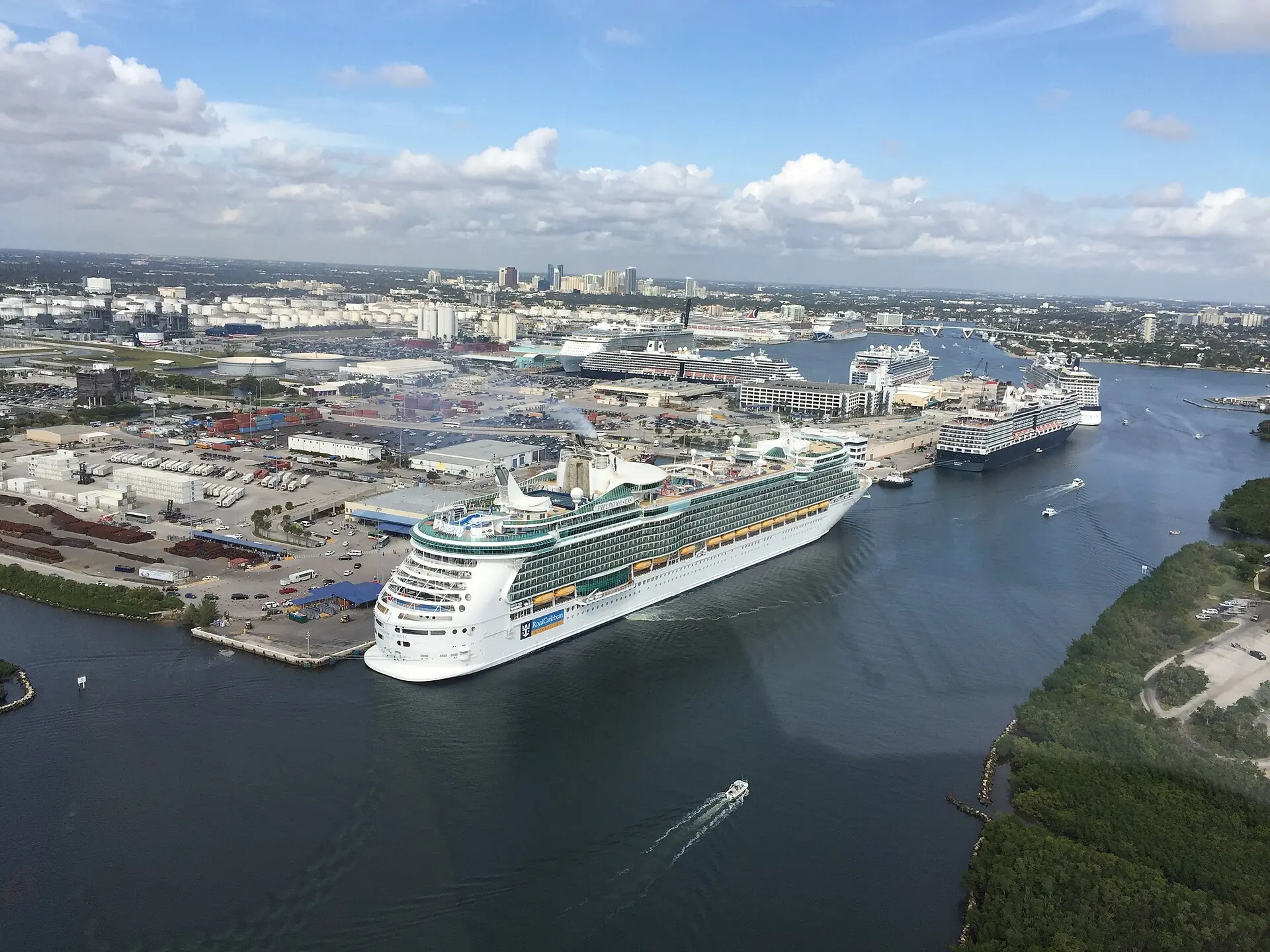 An aerial view of large cruise ships docked and maneuvering through Port Everglades in Fort Lauderdale, with the port facilities and distant skyline visible