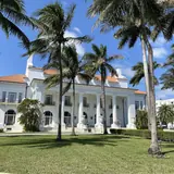 The exterior of the historic Whitehall mansion, the Flagler Museum, framed by tall palm trees across a green lawn in Palm Beach