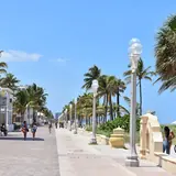 Pedestrians strolling along the paved Hollywood Beach Broadwalk lined with palm trees, classic lampposts, and beachfront architecture