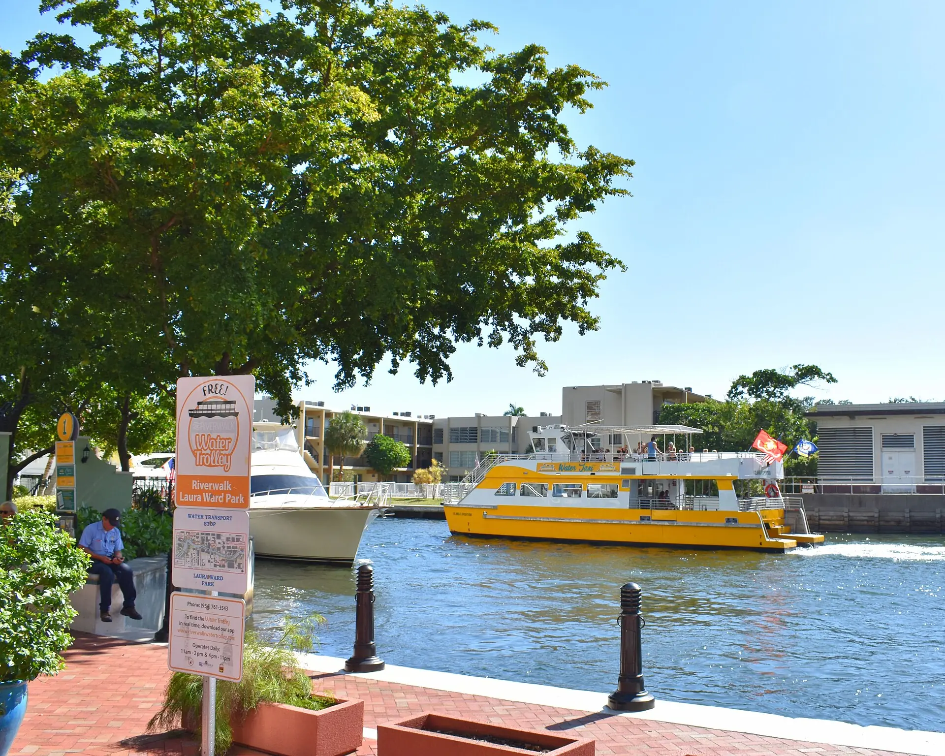 A yellow Fort Lauderdale Water Taxi boat navigates a canal seen from the Riverwalk at Laura Ward Park