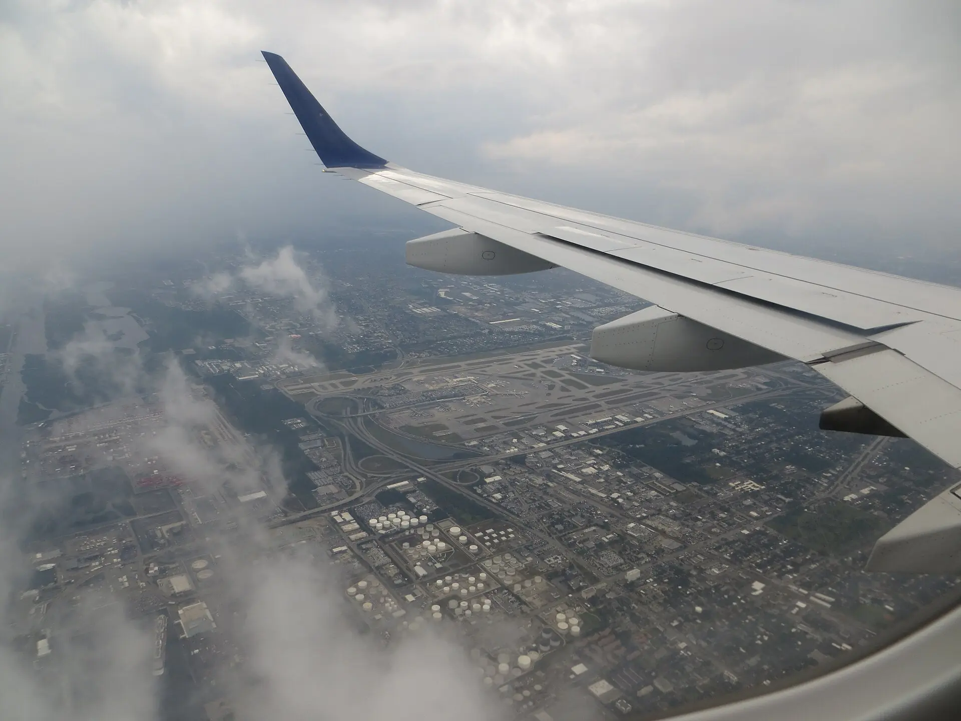 An aerial view through an airplane window showing the aircraft wing above Fort Lauderdale-Hollywood International Airport and Port Everglades under cloudy skies